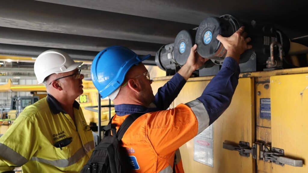 Two workers wearing hard hats (white and blue) inspect a Voltaire pressuriser's filter on an electrical enclosure,